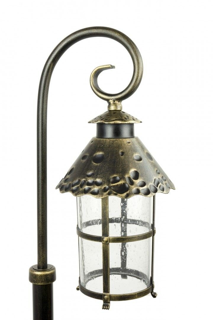 Lampa ogrodowa stojąca z metalowym uchwytem w kolorze patyny mosiądzu, z ozdobnym kloszem z przezroczystego szkła i metalowym daszkiem z fakturą. Klosz jest cylindryczny, otoczony metalowymi prętami.