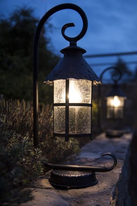 Na zdjęciu widoczna jest stojąca lampa zewnętrzna w kształcie latarni, wykonana z metalu i szkła ozdobnego, emitująca ciepłe światło. Lampa umieszczona jest na kamiennej powierzchni, a w tle widać rozmyte kontury roślinności.