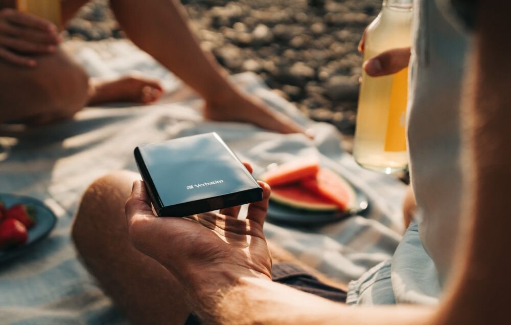 Dysk VERBATIM Store n Go 2TB HDD Czarny Osoba siedząca na kocu trzyma w dłoni czarny dysk przenośny, obok widoczna butelka z napojem i talerz z kawałkiem arbuza, opis