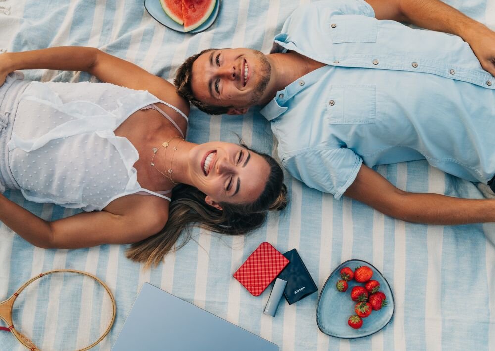 Dysk VERBATIM Store n Go 2TB HDD Czarny Kobieta i mężczyzna leżą na kocu w biało-niebieskie pasy, obok znajdują się dwa dyski przenośne w kolorze czerwonym i czarnym, talerz z truskawkami i kawałkiem arbuza oraz laptop