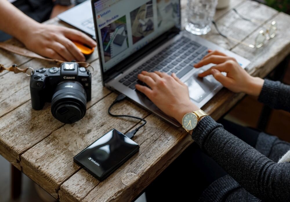 Dysk VERBATIM Store n Go 2TB HDD Czarny Na drewnianym stole stoi aparat fotograficzny, laptop i szklanka z wodą, a obok laptopa podłączony jest czarny dysk przenośny