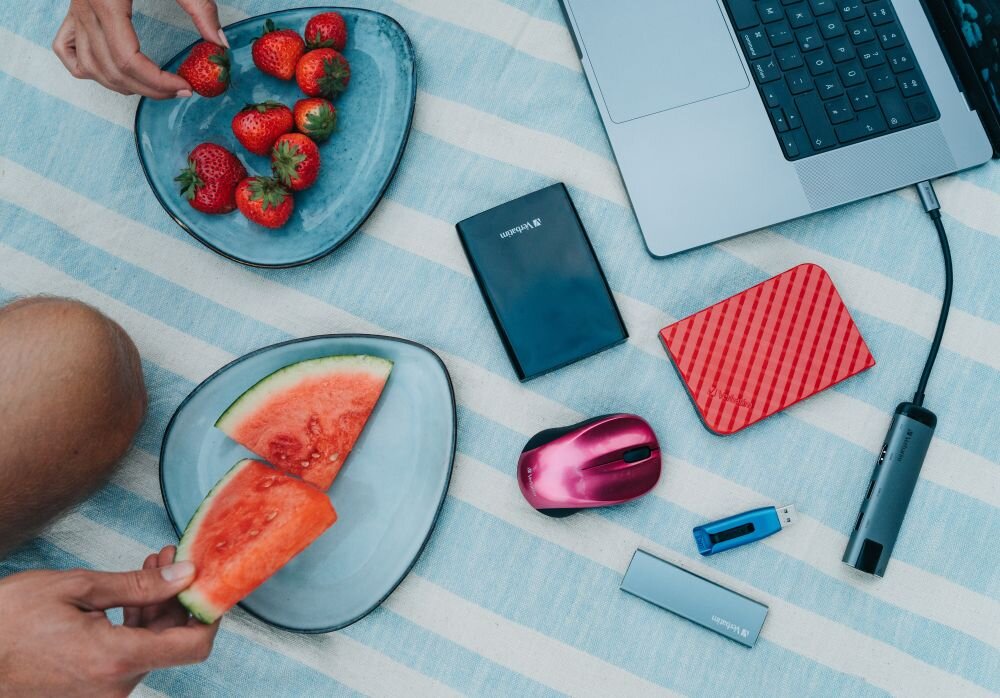 Dysk VERBATIM Store n Go 2TB HDD Czarny Na kocu w biało-niebieskie pasy leżą dwa talerze, jeden z truskawkami, drugi z kawałkami arbuza, obok znajdują się dwa dyski przenośne w kolorze czarnym i czerwonym