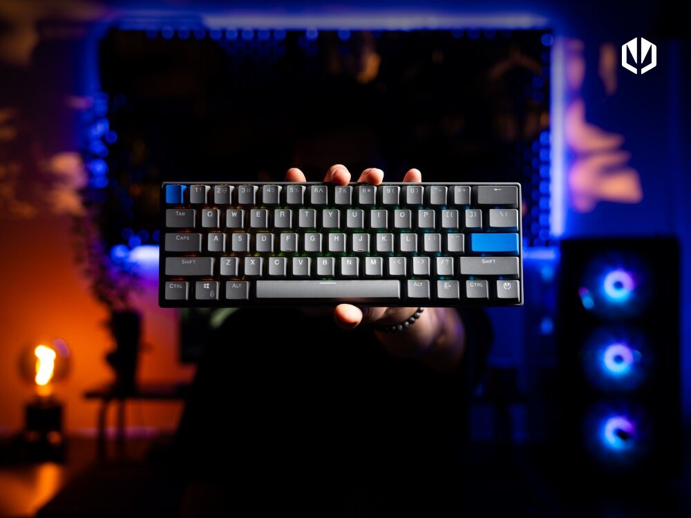 Osoba trzyma klawiaturę mechaniczną z podświetleniem RGB, na której widoczne są klawisze w różnych kolorach. W tle znajduje się rozmyte oświetlenie i komputer z podświetleniem wentylatorów.