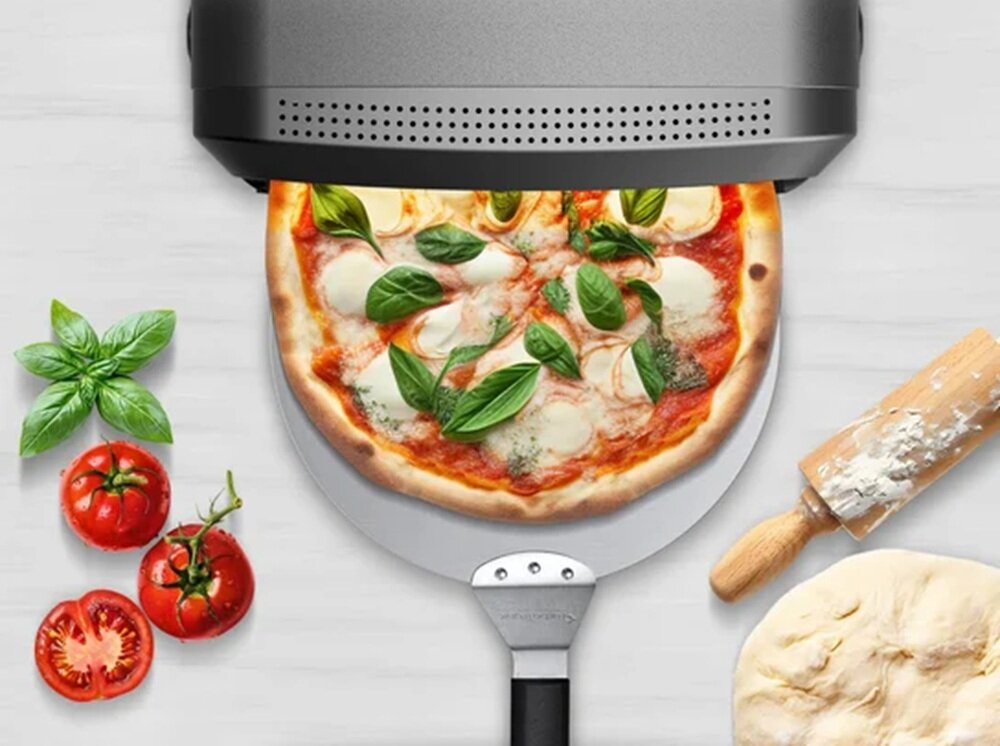 Na obrazie widać piec do pizzy z pizzą na łopatce, obok której znajdują się świeże pomidory, bazylia, wałek do ciasta i kulka ciasta. Piec jest koloru czarnego z perforowanym frontem.