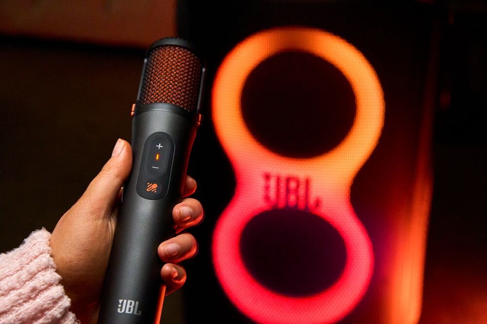 Osoba trzyma mikrofon z logo JBL, w tle widoczny jest głośnik z podświetlanym logo JBL.