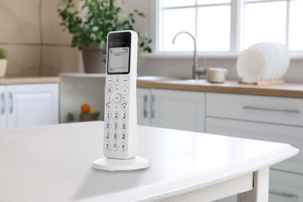 Biały telefon bezprzewodowy stoi na stole w jasnej kuchni. Na ekranie telefonu widoczny jest tekst: \'Handset 1\'.