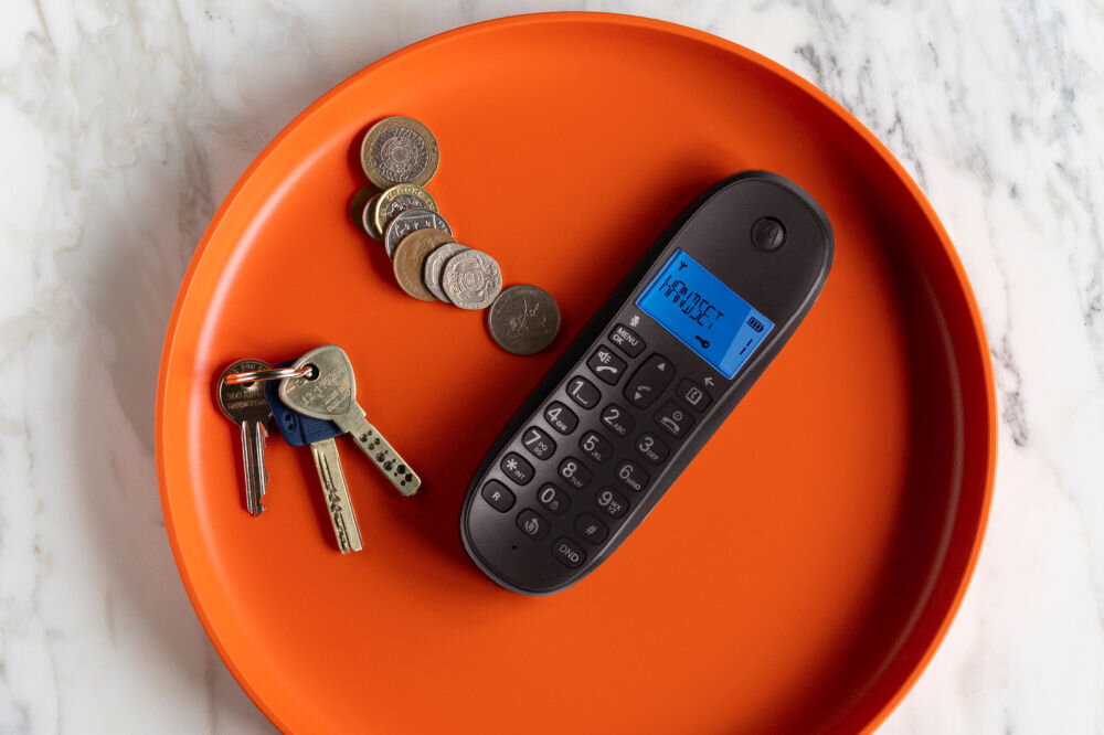 Na pomarańczowej tacy leży czarny telefon z wyświetlaczem pokazującym tekst \'HOUSE\', obok znajdują się klucze i kilka monet. Tło stanowi marmurowa powierzchnia.