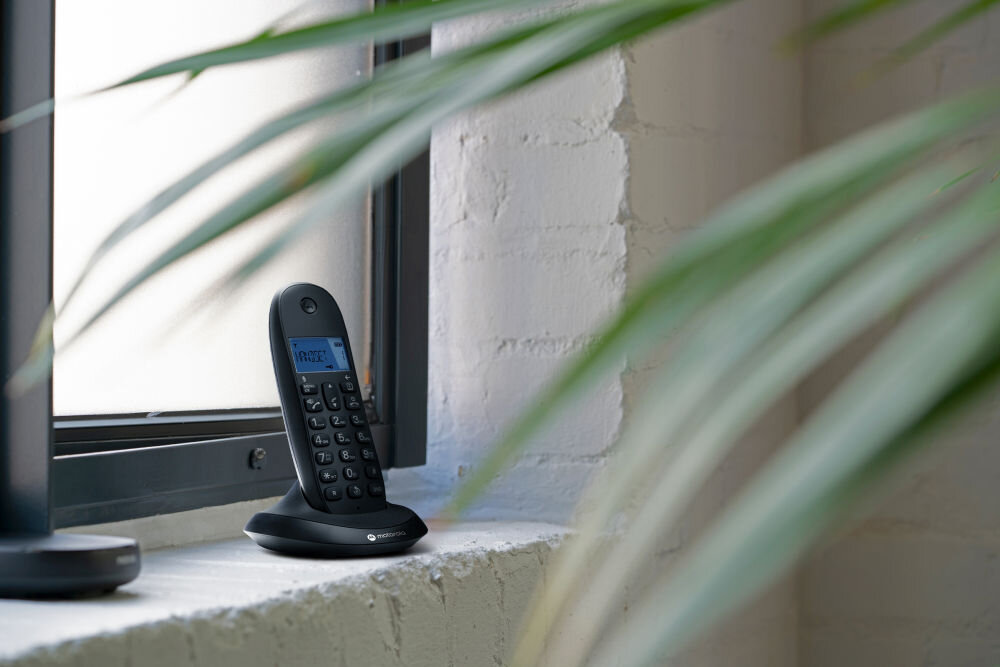 Czarny telefon bezprzewodowy stoi na parapecie przy oknie, z wyświetlaczem pokazującym tekst \'MENU\'.