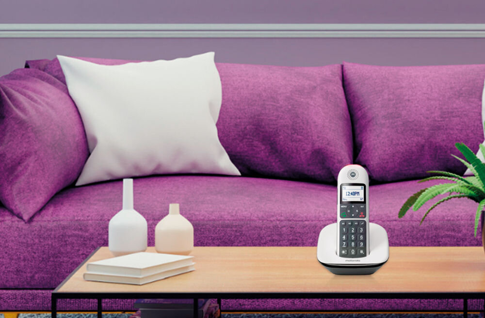 Na drewnianym stoliku stoi biały telefon bezprzewodowy z wyświetlaczem pokazującym godzinę 12:40 PM. W tle znajduje się fioletowa sofa z białą poduszką oraz kilka dekoracyjnych wazonów.