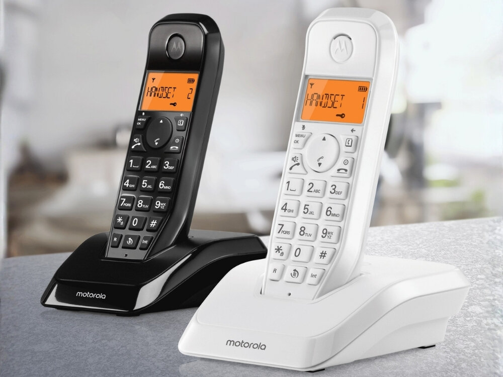 Na obrazie znajdują się dwa bezprzewodowe telefony stacjonarne w kolorach czarnym i białym, każdy z wyświetlaczem pokazującym tekst \'HANDSET\'. Na podstawach telefonów widoczne jest logo \'motorola\'.