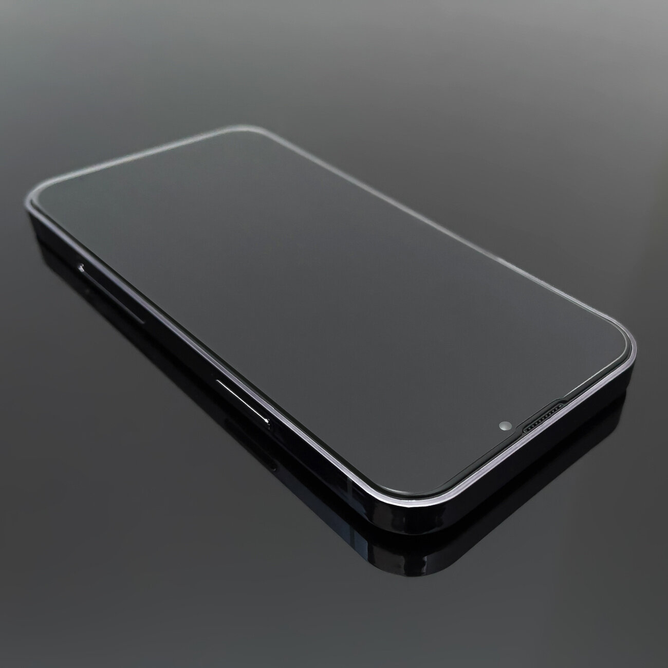 Na obrazie widoczny jest smartfon z nałożonym przezroczystym szkłem ochronnym, leżący na ciemnej, odbijającej powierzchni. Szkło jest gładkie i dopasowane do kształtu ekranu urządzenia.