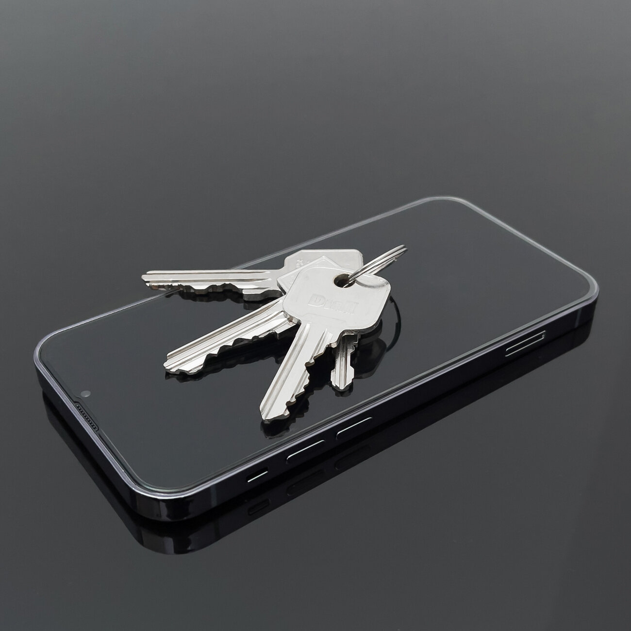 Na ciemnej powierzchni leży smartfon z widocznym szkłem ochronnym, na którym znajdują się trzy metalowe klucze. Telefon ma widoczne przyciski boczne i port ładowania.