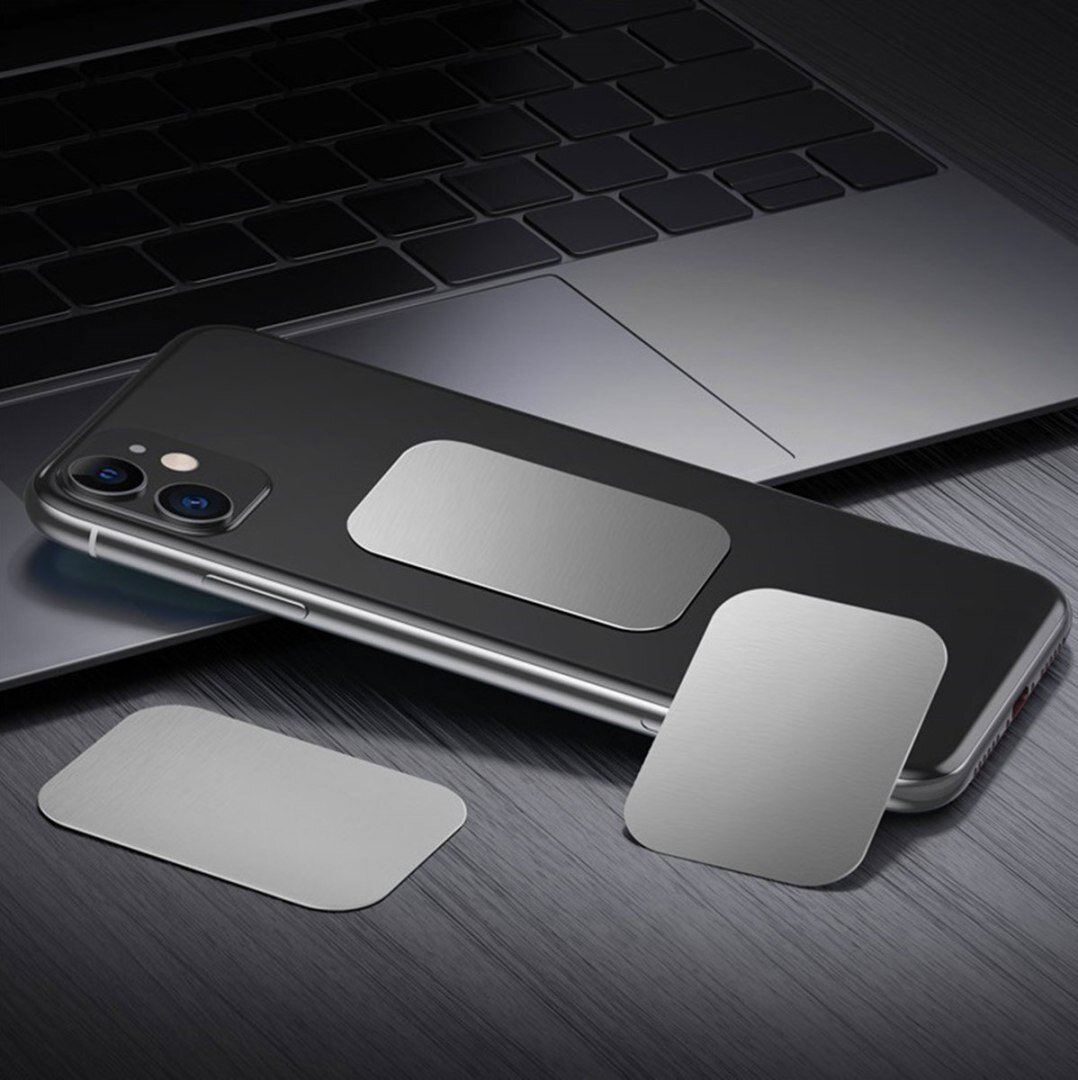 Na obrazie widoczny jest czarny telefon z przyczepioną prostokątną, metaliczną płytką oraz dwie dodatkowe płytki leżące obok na powierzchni przypominającej laptop. Płytki mają srebrny kolor i prostokątny kształt z zaokrąglonymi rogami.