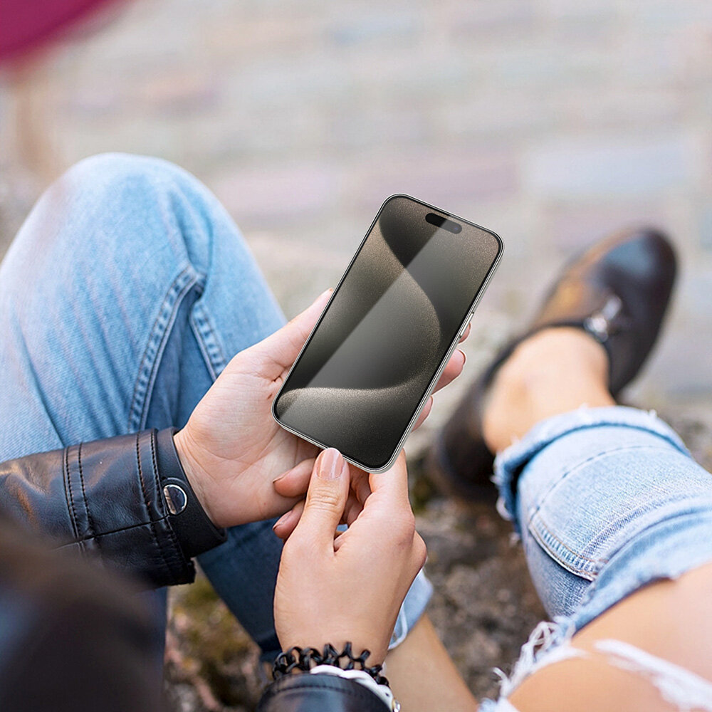 Osoba trzyma smartfon z widocznym ekranem, na którym odbija się światło. W tle widać rozmyty chodnik i fragmenty ubrań osoby.