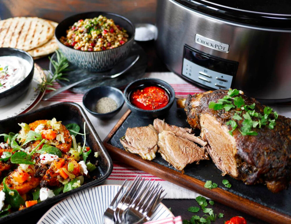 Na stole znajduje się czarno-srebrny wolnowar z widocznym logo \'Crock-Pot\', a obok niego różnorodne potrawy, w tym pieczone mięso i sałatka. W tle widać miski z dodatkami oraz sztućce na talerzu.