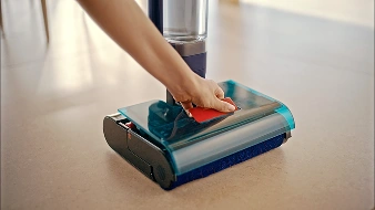Osoba używa elektrycznego mopa z przezroczystą niebieską obudową na gładkiej powierzchni. Mop ma widoczny czerwony element na górze.