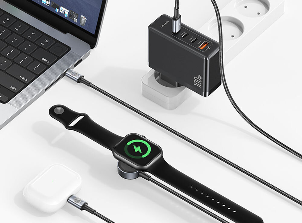 Na obrazie znajduje się czarna ładowarka z kilkoma portami USB, podłączona do listwy zasilającej. Obok niej leży smartwatch na ładowarce indukcyjnej oraz laptop z podłączonym kablem USB-C.