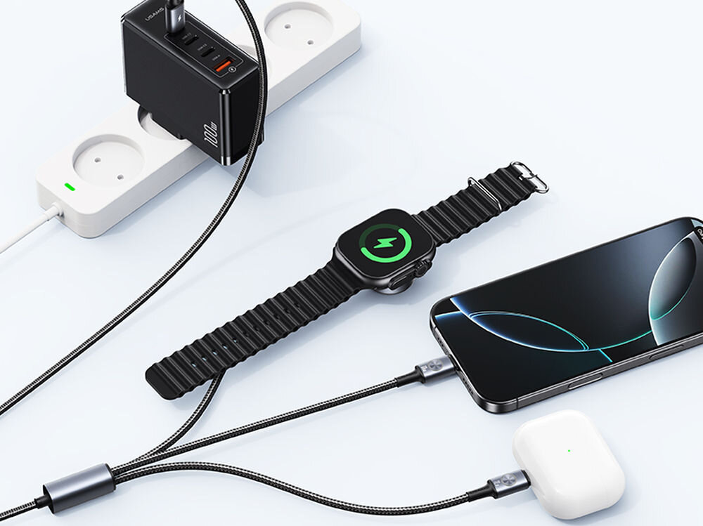 Na obrazie widoczna jest czarna ładowarka z kilkoma portami USB podłączona do listwy zasilającej, obok której znajdują się smartwatch, smartfon i etui na słuchawki, wszystkie podłączone do ładowania. Na ładowarce widoczny jest napis \'100W\'.