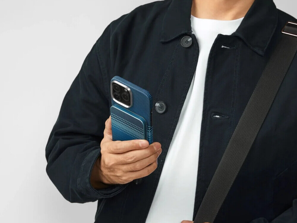 Osoba trzyma niebieski powerbank przymocowany do telefonu, ubrana w ciemną kurtkę i białą koszulkę. Powerbank ma teksturowaną powierzchnię i jest dopasowany kolorystycznie do etui telefonu.
