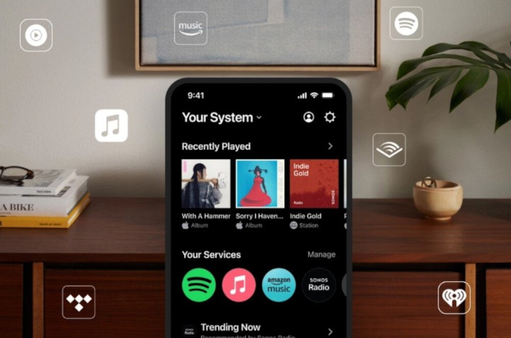 Na obrazie widać smartfon z aplikacją muzyczną, na której ekranie wyświetlane są albumy i usługi muzyczne takie jak Spotify, Apple Music i Amazon Music. W tle znajduje się drewniana komoda z książkami i rośliną.