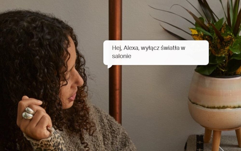Osoba siedzi obok rośliny w doniczce, a na obrazie widoczny jest tekst: \'Hej, Alexa, wyłącz światła w salonie\'.