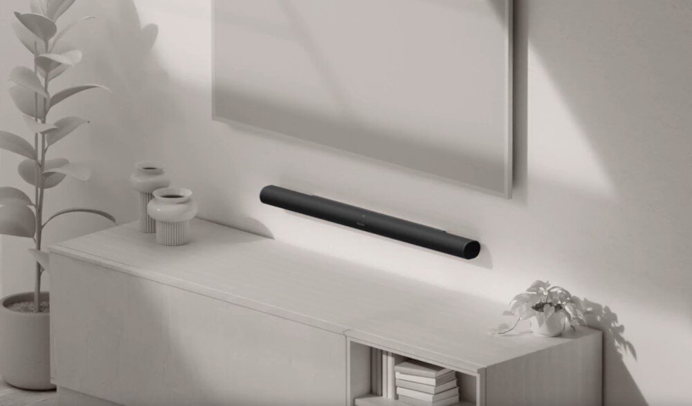 Czarny soundbar zamontowany na ścianie pod dużym ekranem, obok jasnej szafki z dekoracyjnymi wazonami i rośliną.