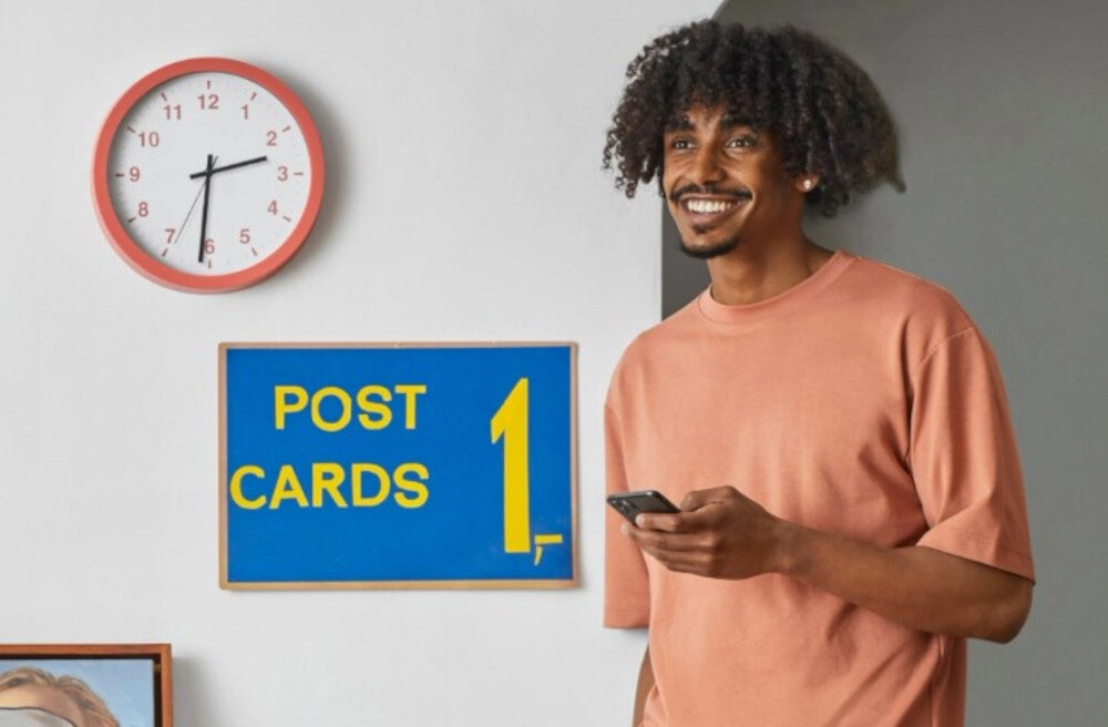 Na ścianie wisi zegar z czerwonym obramowaniem oraz tablica z napisem \'POST CARDS 1,-\'. Obok stoi osoba trzymająca telefon.