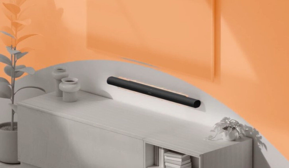 Na obrazie widać czarny soundbar zamontowany na ścianie nad jasną szafką. W tle znajduje się pomarańczowa ściana, a na szafce stoją dwa dekoracyjne naczynia i roślina.