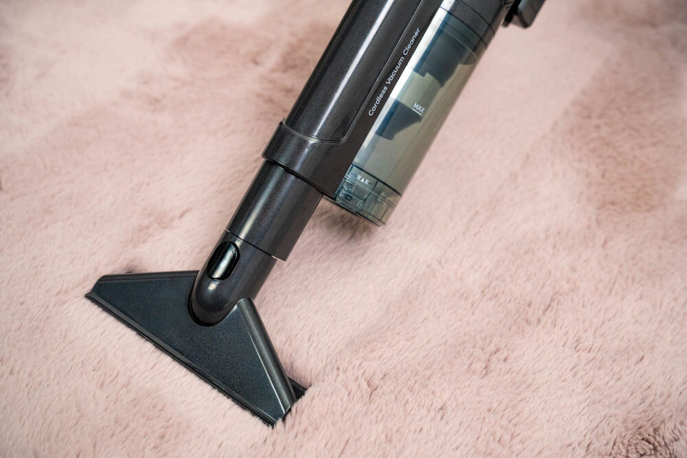 Odkurzacz ręczny z przezroczystym pojemnikiem na kurz, widoczny na różowym dywanie. Na urządzeniu znajduje się napis \'Cordless Vacuum Cleaner\'.