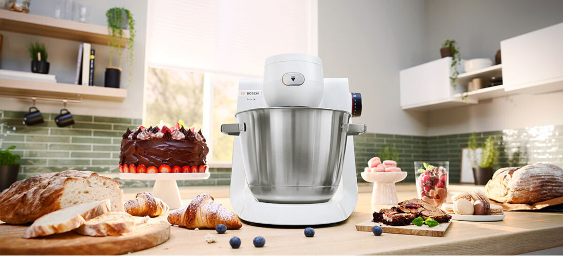 Na blacie kuchennym stoi biały robot kuchenny z metalową misą. W tle widoczne są wypieki, owoce i dekoracje kuchenne.