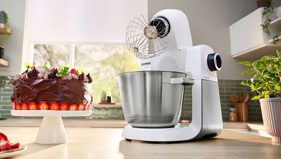 Na blacie kuchennym stoi biały robot kuchenny z metalową misą i trzepaczką. Obok znajduje się tort czekoladowy ozdobiony truskawkami i zielonymi liśćmi.