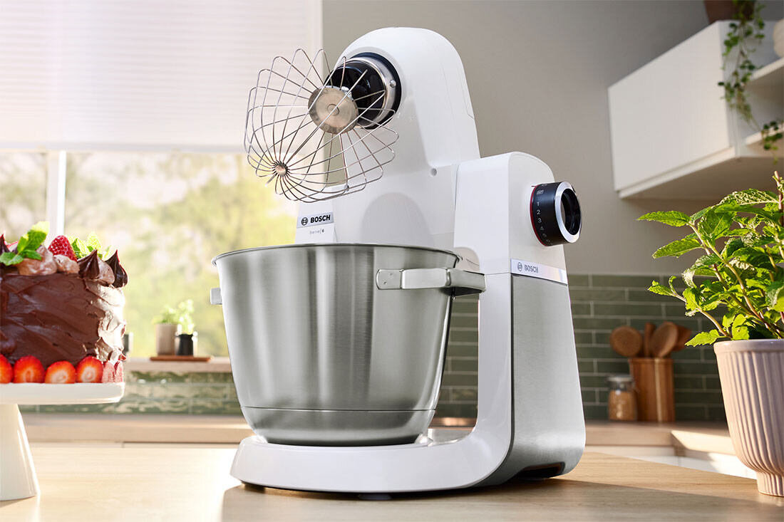 Na blacie kuchennym stoi biały robot kuchenny z metalową misą i trzepaczką. Obok znajduje się tort czekoladowy ozdobiony truskawkami, a w tle widać rośliny doniczkowe i kuchenne akcesoria.