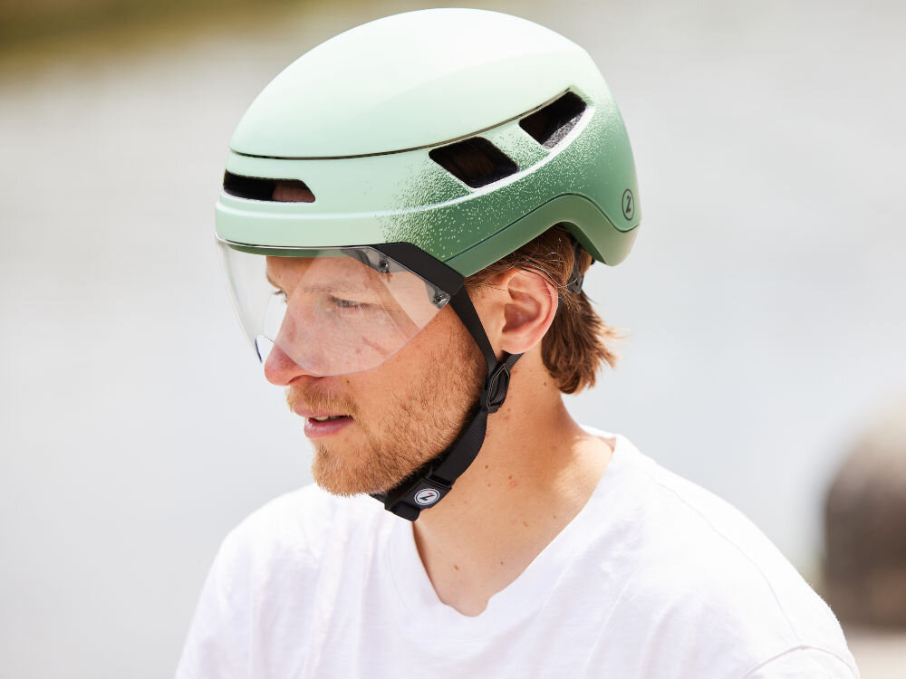 Osoba nosi zielony kask rowerowy z przezroczystą osłoną i otworami wentylacyjnymi. Kask ma widoczne logo w formie litery \'Z\'.