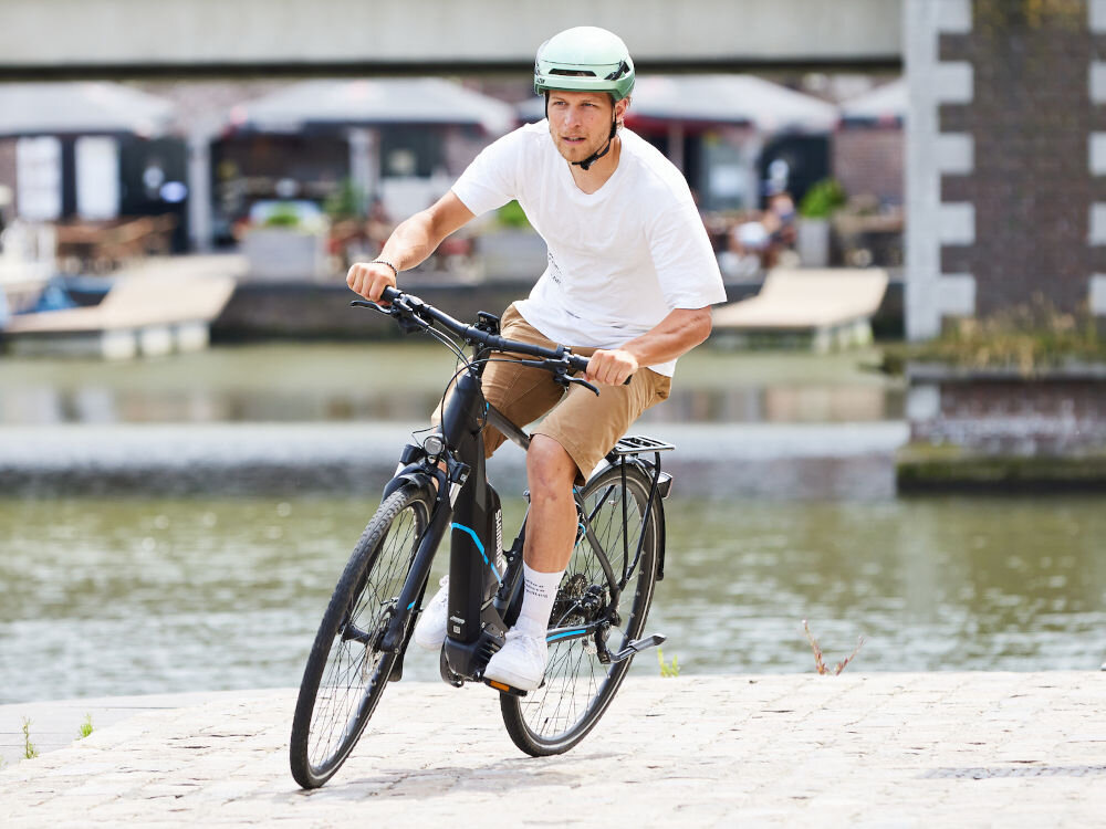 Osoba na rowerze nosi zielony kask rowerowy. W tle widać wodę i zabudowania miejskie.