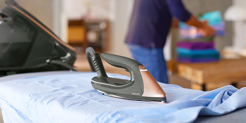 Żelazko z napisem \'PHILIPS\' stoi na desce do prasowania pokrytej niebieskim materiałem. W tle osoba układa kolorowe ubrania na stole.