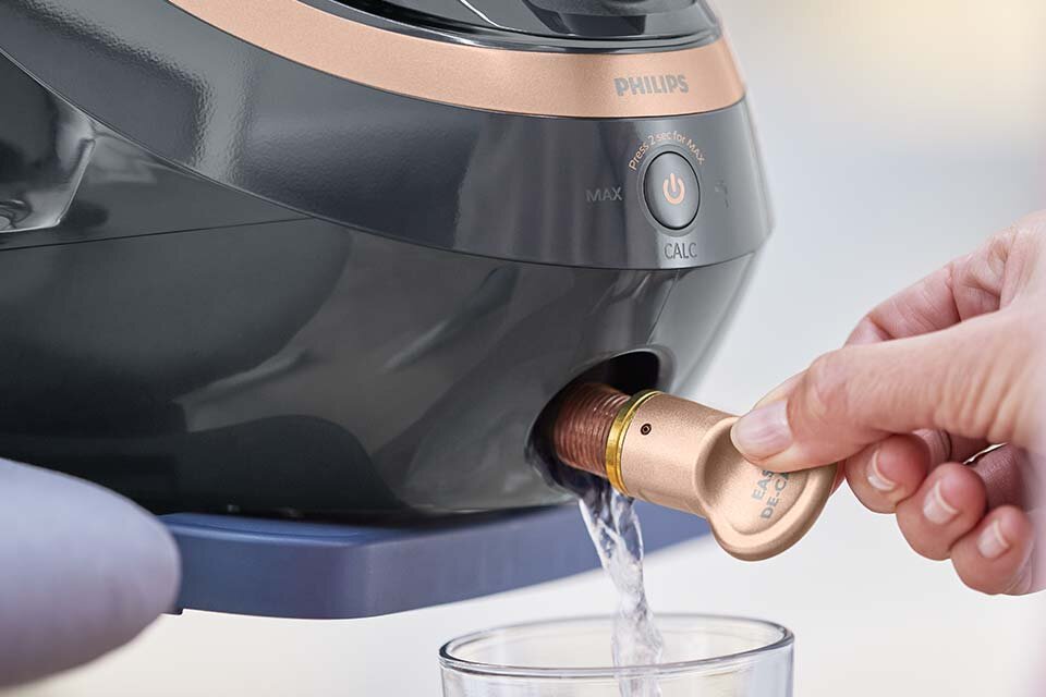 Osoba trzyma złoty element urządzenia, z którego wypływa woda do szklanki. Na urządzeniu widoczne jest logo \'PHILIPS\' oraz przycisk z napisem \'CALC\'.