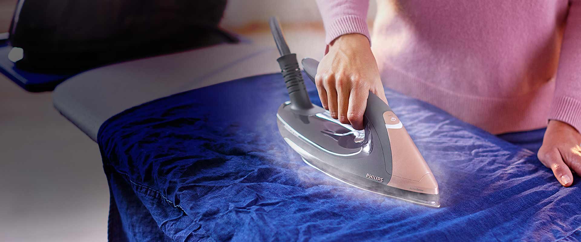 Osoba prasuje niebieski materiał za pomocą żelazka z widocznym logo Philips. Żelazko emituje parę podczas prasowania.