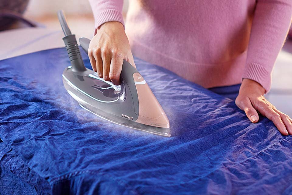 Osoba prasuje niebieski materiał za pomocą żelazka parowego z widocznym logo PHILIPS. Z żelazka wydobywa się para, a osoba nosi różowy sweter.