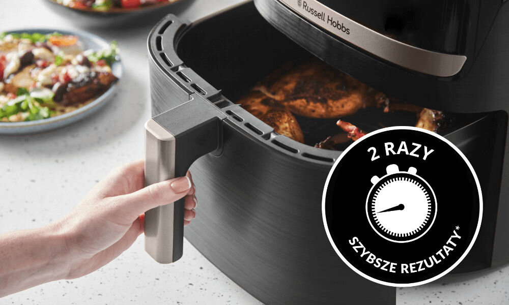 Osoba trzyma uchwyt czarnej frytkownicy beztłuszczowej marki Russell Hobbs, w której widać przygotowywane jedzenie. Na obrazie znajduje się także okrągły symbol z tekstem \'2 RAZY SZYBSZE REZULTATY\'.