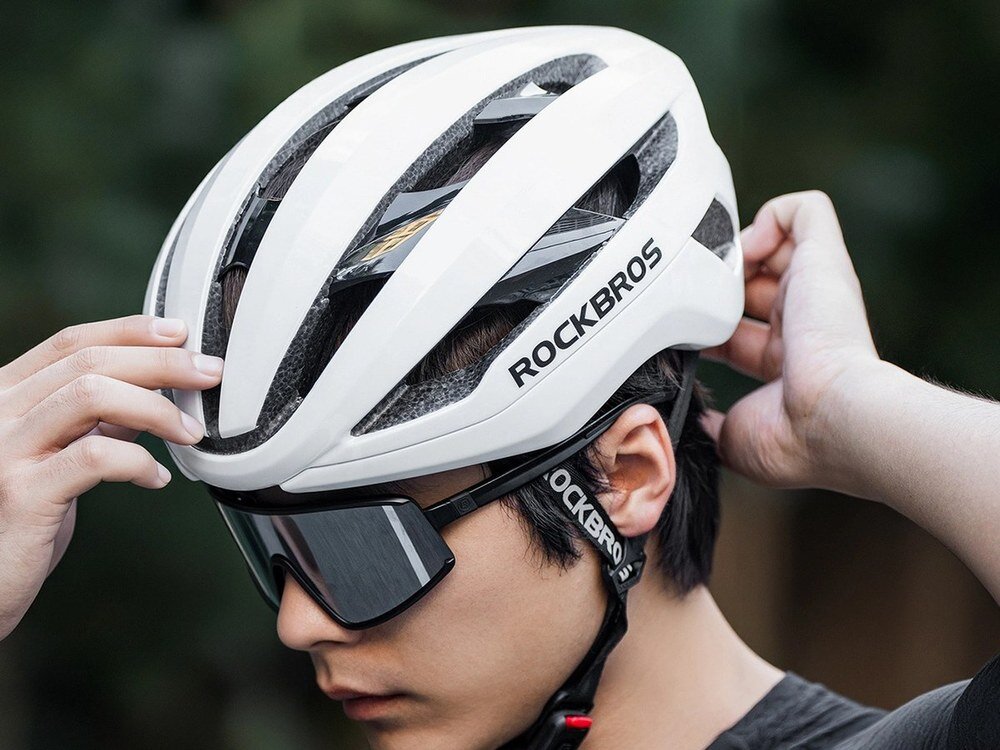 Osoba zakłada biały kask rowerowy z napisem ROCKBROS. Kask ma aerodynamiczny kształt i widoczne otwory wentylacyjne.