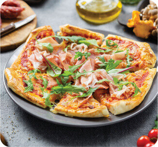Na talerzu znajduje się pizza z szynką i rukolą. W tle widoczne są składniki takie jak oliwa i grzyby.