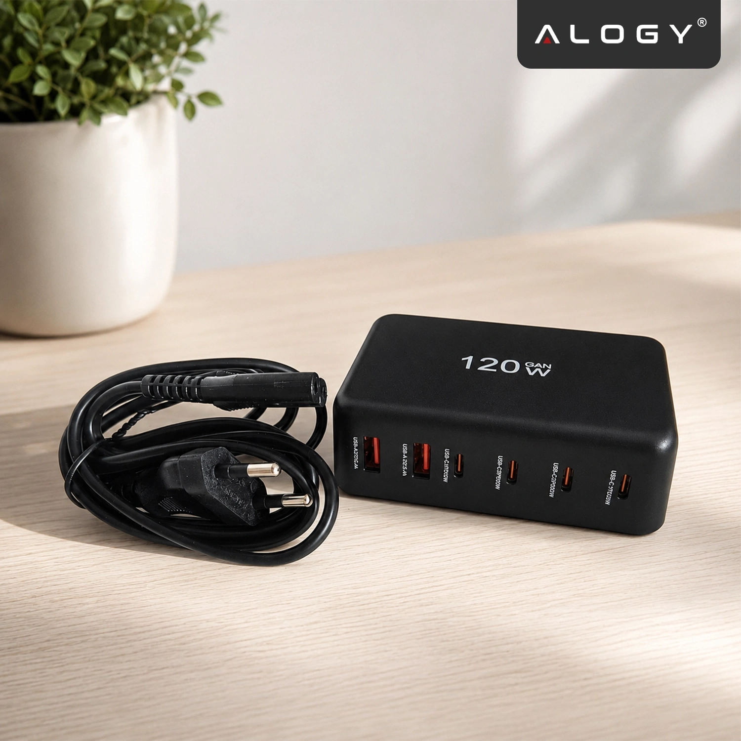 Czarna ładowarka biurkowa z sześcioma portami (4x USB-C, 2x USB-A) i napisem \'120W GAN\' na górze, obok zwiniętego kabla z wtyczką. W tle widoczna jest roślina w doniczce, a w prawym górnym rogu logo \'ALOGY\'.