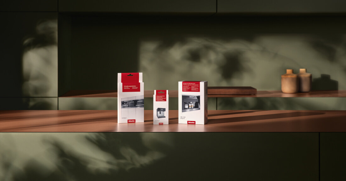 Na drewnianym blacie stoją trzy pudełka z produktami Miele, każde z czerwonym logo Miele na dole. W tle widać zieloną ścianę z cieniami roślin.