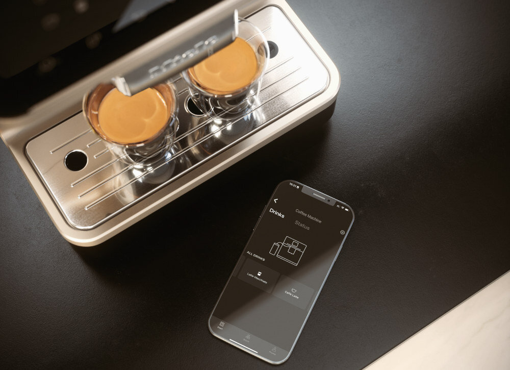 Na blacie znajduje się ekspres do kawy z dwoma filiżankami wypełnionymi kawą oraz smartfon z aplikacją do obsługi ekspresu. Na ekranie telefonu widoczny jest tekst: \'Drinks\', \'Coffee Machine\', \'Settings\'.