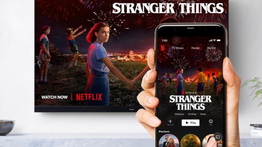 Na ekranie telewizora wyświetlany jest plakat serialu \'Stranger Things\' z logo Netflix i napisem \'WATCH NOW\'. Przed telewizorem osoba trzyma smartfon z aplikacją Netflix, na której również widoczny jest plakat \'Stranger Things\'.