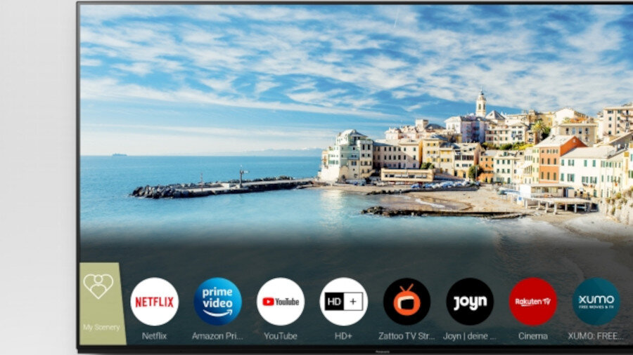 Ekran telewizora wyświetla obraz nadmorskiego miasteczka z aplikacjami na dole, w tym Netflix, Amazon Prime Video, YouTube i inne. Widoczne są ikony aplikacji oraz teksty: \'My Scenery\', \'Netflix\', \'Amazon Prime Video\', \'YouTube\', \'HD+\', \'Zattoo TV Streaming\', \'Joyn | deine Streaming App\', \'Rakuten TV\', \'Cinema\', \'XUMO: FREE\'.