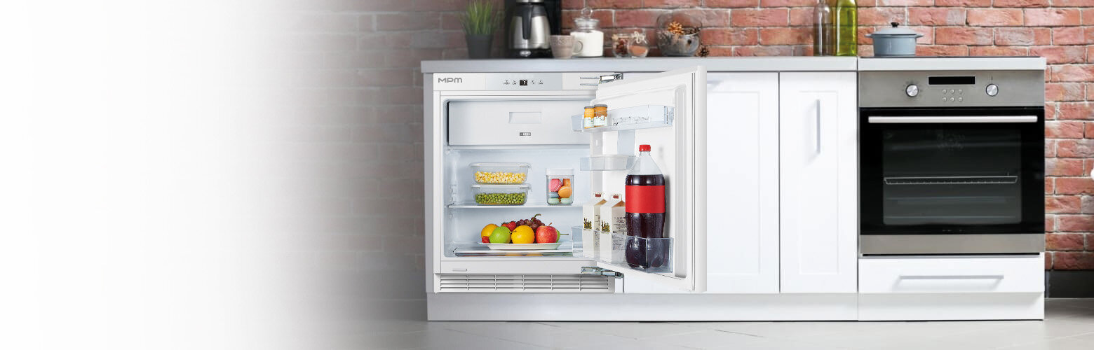 Kuchnia z otwartą lodówką w zabudowie, w której znajdują się owoce, napoje i produkty spożywcze. Obok lodówki znajduje się piekarnik, a na blacie stoją różne przedmioty kuchenne.