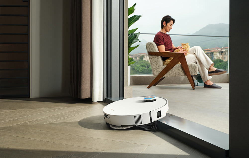 Biały robot sprzątający porusza się po podłodze w nowoczesnym wnętrzu z dużymi oknami. W tle widać osobę siedzącą na krześle z kotem na kolanach.