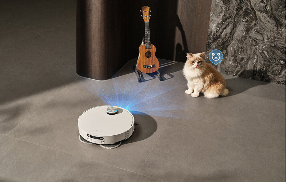 Na podłodze znajduje się biały robot sprzątający, obok którego stoi gitara i siedzi kot. Robot emituje niebieskie światło w kierunku podłogi.