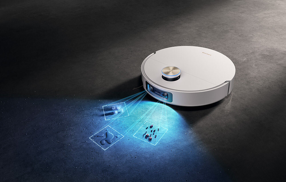 Biały robot sprzątający na ciemnej podłodze, emitujący niebieskie światło skanujące powierzchnię. Na obudowie widoczne jest logo producenta.